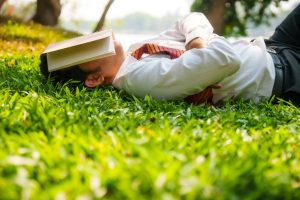 Asian businessman lying down on grass area and napping with arms crossed and book covering his face to protect eyes from sunlight during his lunch break at a park. MSP bedtime stories